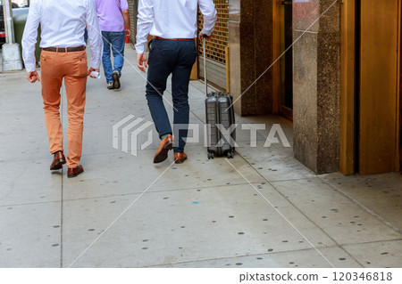 Two men in dress shirts, slacks stroll along busy city sidewalk, one pulling carry on suitcase, indicating travel professional meeting nearby. 120346818