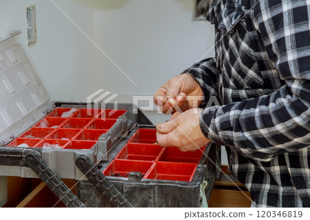 Man sorts through small components in toolbox with red containers, focusing on organizing materials at workshop setting. Man sorts through small components in toolbox with red containers, focusing on organizing materials at workshop setting. 120346819