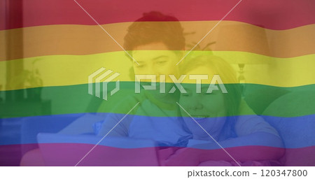 Image of pride rainbow flag over happy caucasian lesbian couple using laptop in bed 120347800