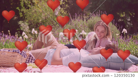 Image of red heart balloons over happy diverse couple relaxing at picnic in sunny garden 120350130