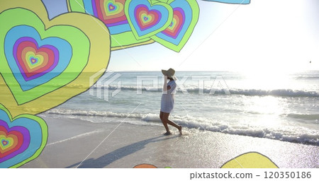 Image of colourful hearts over biracial woman walking on beach 120350186