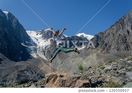 Young woman in a jump against the backdrop of the high mountains of Altai with snow-capped peaks and a glacier. 120350937