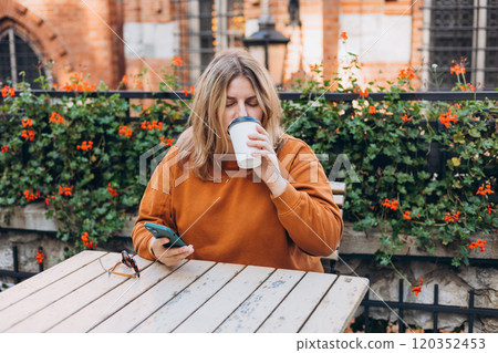 Woman holding bio coffee paper cup and smartphone, she is sitting on the cafe terrace on the city street. Urban concept. Happy Girl sitting at a table with a coffee using mobile phone. 120352453