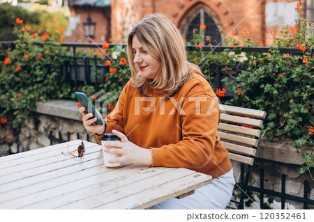 Woman holding bio coffee paper cup and smartphone, she is sitting on the cafe terrace on the city street. Urban concept. Happy Girl sitting at a table with a coffee using mobile phone. Woman holding bio coffee paper cup and smartphone, she is sitting on the cafe terrace on the city street. Urban concept. Happy Girl sitting at a table with a coffee using mobile phone. 120352461