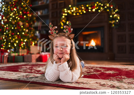 Merry Christmas. Little girl smiling near Christmas tree in classical dark interior. Young happy kid wearing deer horns in living room with fireplace Christmas tree gift boxes. Christmas eve at home Merry Christmas. Little girl smiling near Christmas tree in classical dark interior. Young happy kid wearing deer horns in living room with fireplace Christmas tree gift boxes. Christmas eve at home 120353327