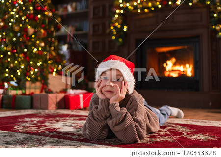 Merry Christmas. Little boy smiling near Christmas tree in classical dark interior. Young happy kid wearing Santa Hat in living room with fireplace Christmas tree gift boxes. Christmas eve at home Merry Christmas. Little boy smiling near Christmas tree in classical dark interior. Young happy kid wearing Santa Hat in living room with fireplace Christmas tree gift boxes. Christmas eve at home 120353328