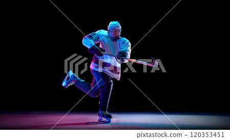 Competitive man, hockey player wearing full game gear, in motion with stick, competing for the victory against black studio background in neon light 120353451