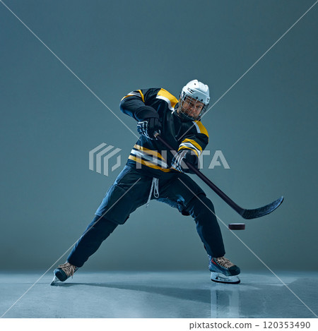 Hockey player wearing black and yellow uniform, captured in mid-action with stick to hit puck, against studio background. 120353490