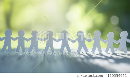 A group of connected paper figures holding hands on a wooden surface under soft sunlight in a natural setting 120354651