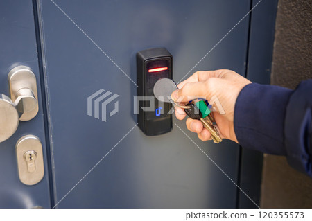 Close up view of person using a electric lock key fob to access a building via a reader of entry system mounted on a house wall 120355573