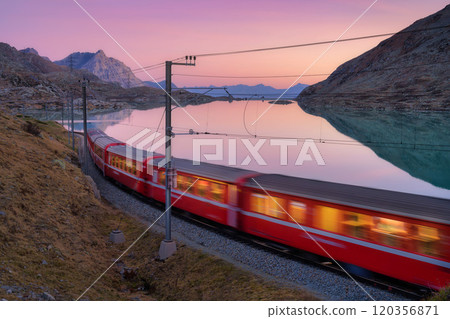 Moving red train near azure mountain lake, snowy rocks at sunset 120356871