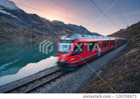Moving red train near azure mountain lake, snowy rocks at sunset 120356873