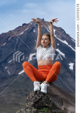 Fitness instructor sits on stone with her eyes closed and arms raised, stretching and warming up before climbing high cone of volcano. Millennial blonde woman enjoying moment of summer sports training 120358178
