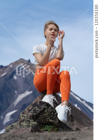 Woman athlete sits on rocky outcrop in mountains, with magnificent cone of volcano looming in distance. Millennial woman enjoying moment of repose, raises her hands to face and gazes into distance Woman athlete sits on rocky outcrop in mountains, with magnificent cone of volcano looming in distance. Millennial woman enjoying moment of repose, raises her hands to face and gazes into distance 120358180