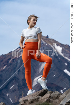Millennial Caucasian woman stands proudly on rocky mountaintop and looks into distance against backdrop of volcano, blue sky. Adult woman yoga instructor in T-shirt, orange yoga leggings and trainers Millennial Caucasian woman stands proudly on rocky mountaintop and looks into distance against backdrop of volcano, blue sky. Adult woman yoga instructor in T-shirt, orange yoga leggings and trainers 120358189