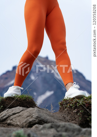Unrecognizable female athlete training in mountains with volcano in background. She's wearing orange skintight workout leggings and sneakers. Close-up low section of female body during outdoor workout 120358208