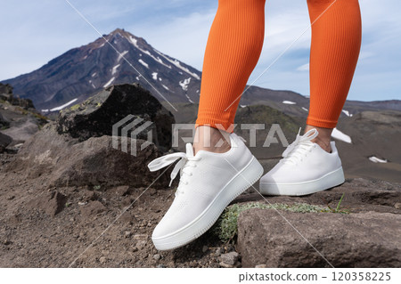 Close-up view of woman's legs wearing white trainers and orange sports leggings. Woman stands during fitness workout in mountains with beautiful view of volcano cone on summer day Close-up view of woman's legs wearing white trainers and orange sports leggings. Woman stands during fitness workout in mountains with beautiful view of volcano cone on summer day 120358225