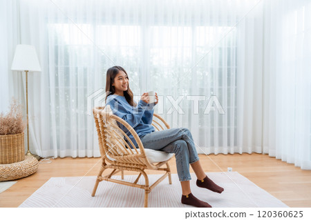 Young Asian woman happy and smiling, sitting on seat and holding mug in living room at home 120360625