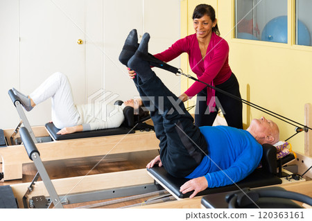 Pilates instructor helping older man do leg stretch on the machine in gym 120363161