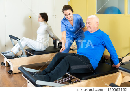 Medical professional helps and monitors the difficulty of Pilates exercise for an mature man Medical professional helps and monitors the difficulty of Pilates exercise for an mature man 120363162
