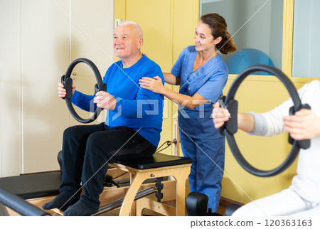Rehabilitation doctor helping elderly man exercising with pilates ring 120363163