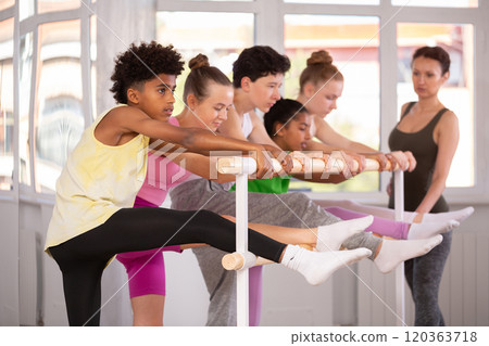 Teenage boy doing leg stretching exercise holding by ballet barre during dance courses 120363718