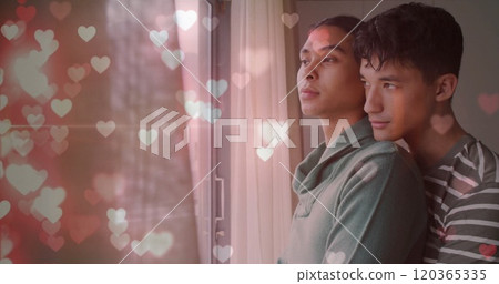 Image of glittering hearts over happy diverse gay male couple looking out of window, embracing Image of glittering hearts over happy diverse gay male couple looking out of window, embracing 120365335