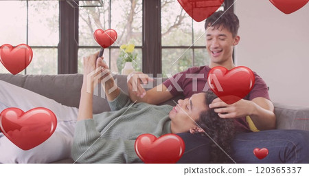 Image of red hearts over happy diverse gay male couple relaxing on couch using smartphone Image of red hearts over happy diverse gay male couple relaxing on couch using smartphone 120365337