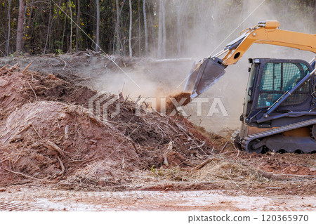 Construction bulldozer machinery is actively clearing dirt debris in forested location dust rises as excavator scoops up materials, preparing for future development. 120365970