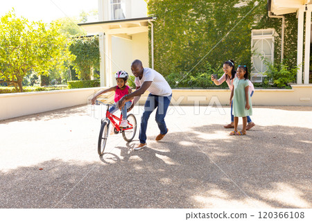 Teaching child to ride bicycle, father supporting while mother and sister watching 120366108