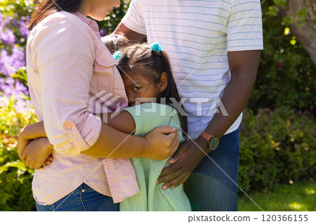 Hugging parents, young girl looking shy in family bonding moment outdoors 120366155