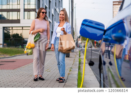 Two women stand by a charging station holding bags with grocery, while electric car charging on the parking lot  120366814