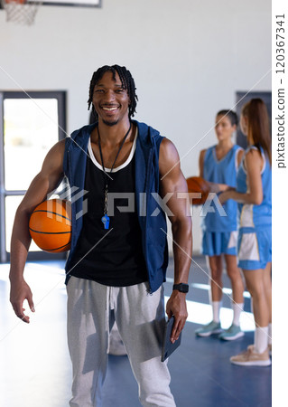 male african American basketball coach holding ball and whistle, smiling in gym with female players 120367341