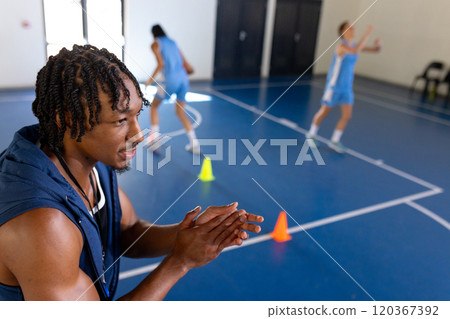 Encouraging female players, male african american basketball coach clapping at practice in gym 120367392