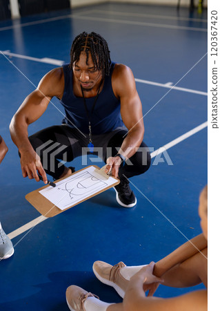 Male coach explaining basketball strategy on clipboard to female players in gym Male coach explaining basketball strategy on clipboard to female players in gym 120367420