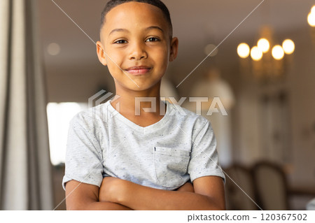 Smiling boy standing with arms crossed in living room, feeling confident 120367502