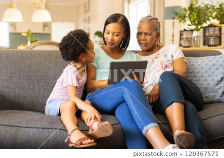 Using tablet, multi-generational family sitting on couch, engaging in conversation 120367571