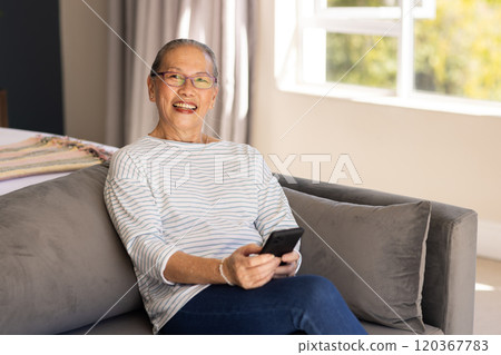 Smiling elderly Asian woman sitting on couch holding smartphone in living room 120367783