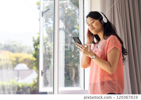 Listening to music on headphones, Asian woman enjoying smartphone by window, copy space 120367819