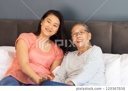 Smiling Asian grandmother and granddaughter sitting together on bed, enjoying time 120367838