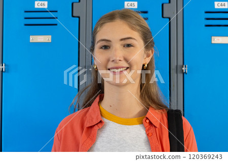 Smiling teenage girl standing in front of blue lockers at school, carrying backpack 120369243