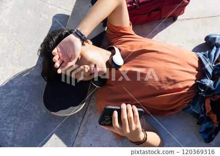 Resting on skateboard, asian teenage boy using smartphone and wearing headphones outdoors 120369316