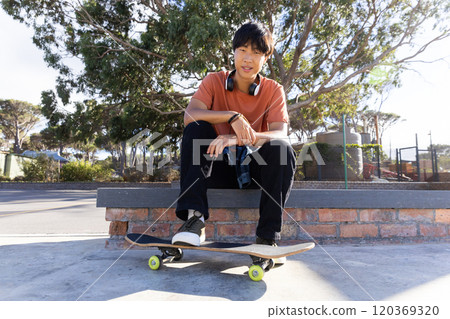 Sitting on skateboard, asian teenage boy with headphones relaxing outdoors in park 120369320