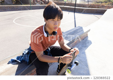 Outdoors, Sitting on bench, asian teenage boy holding skateboard and wearing headphones at skatepark 120369322