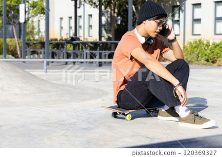 Outdoors, Sitting on skateboard, asian teenage boy talking on smartphone and wearing headphones Outdoors, Sitting on skateboard, asian teenage boy talking on smartphone and wearing headphones 120369327
