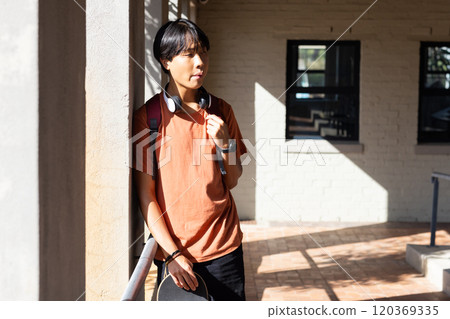 At school, Asian teenage boy with headphones leaning on railing, holding skateboard, enjoying break 120369335
