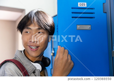 Opening locker, smiling asian teenage boy with braces and headphones in school hallway Opening locker, smiling asian teenage boy with braces and headphones in school hallway 120369350