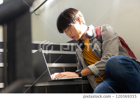 At school, Asian teenage boy sitting on stairs using laptop with headphones around neck At school, Asian teenage boy sitting on stairs using laptop with headphones around neck 120369355