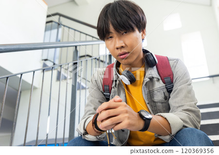 At school, Asian teenage boy with backpack and headphones sitting on stairs, looking thoughtful 120369356