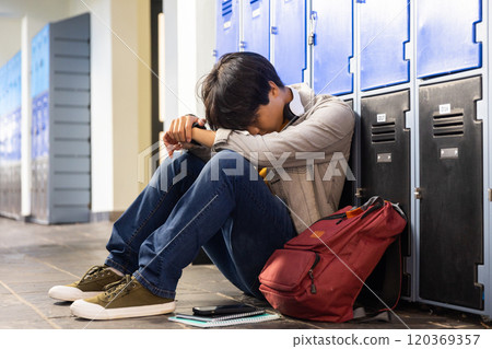 Sitting by school lockers, asian teenage boy resting head on arms with backpack and notebook 120369357